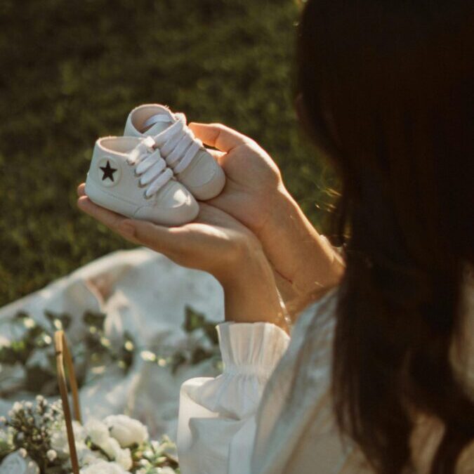 A woman lovingly holds baby shoes in a tranquil garden setting with white roses.